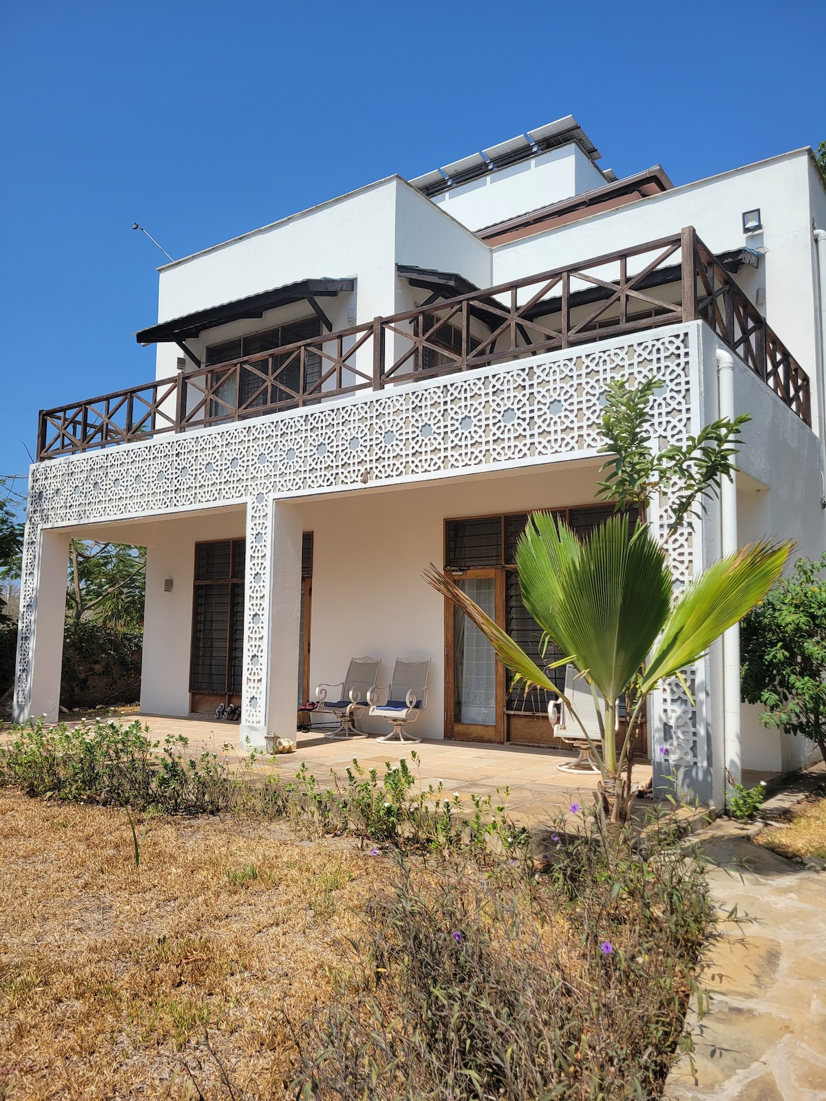 A two-story house features a decorative white facade with intricate patterns. A spacious patio is furnished with two lounge chairs, set against a backdrop of well-kept plants and a clear blue sky. Sunlight highlights the natural tones of the surrounding area.