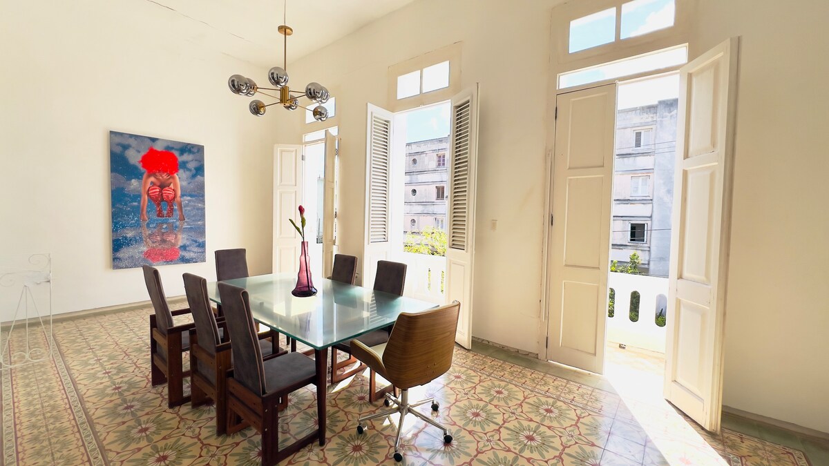 A bright dining area is showcased with a large glass table surrounded by eight chairs. Natural light enters through multiple open windows, highlighting the artwork on the wall. The tiled floor features intricate patterns, creating a vibrant contrast with the light-colored walls.