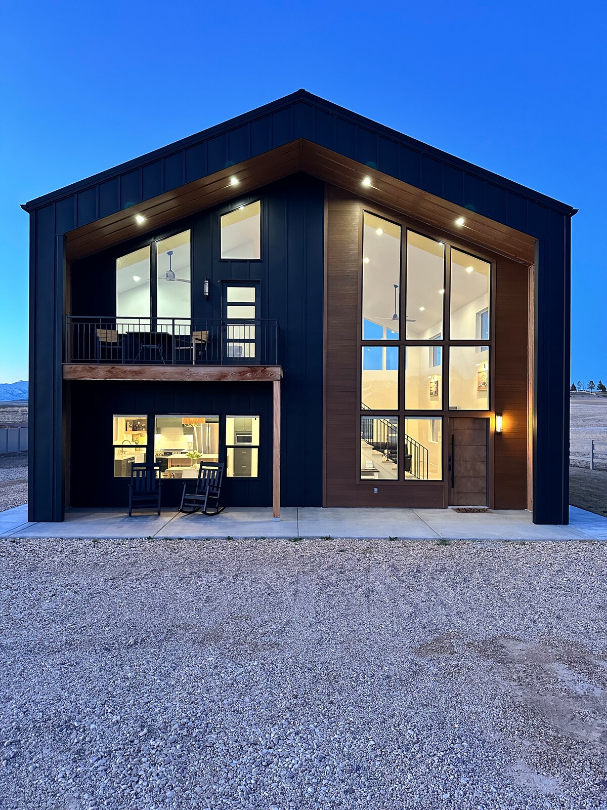 The exterior of the farmhouse reveals a modern two-story design, featuring large glass windows and illuminated spaces. A porch with rocking chairs is positioned on the upper level, while the entrance is accessed through a sturdy door framed by warm wood accents.