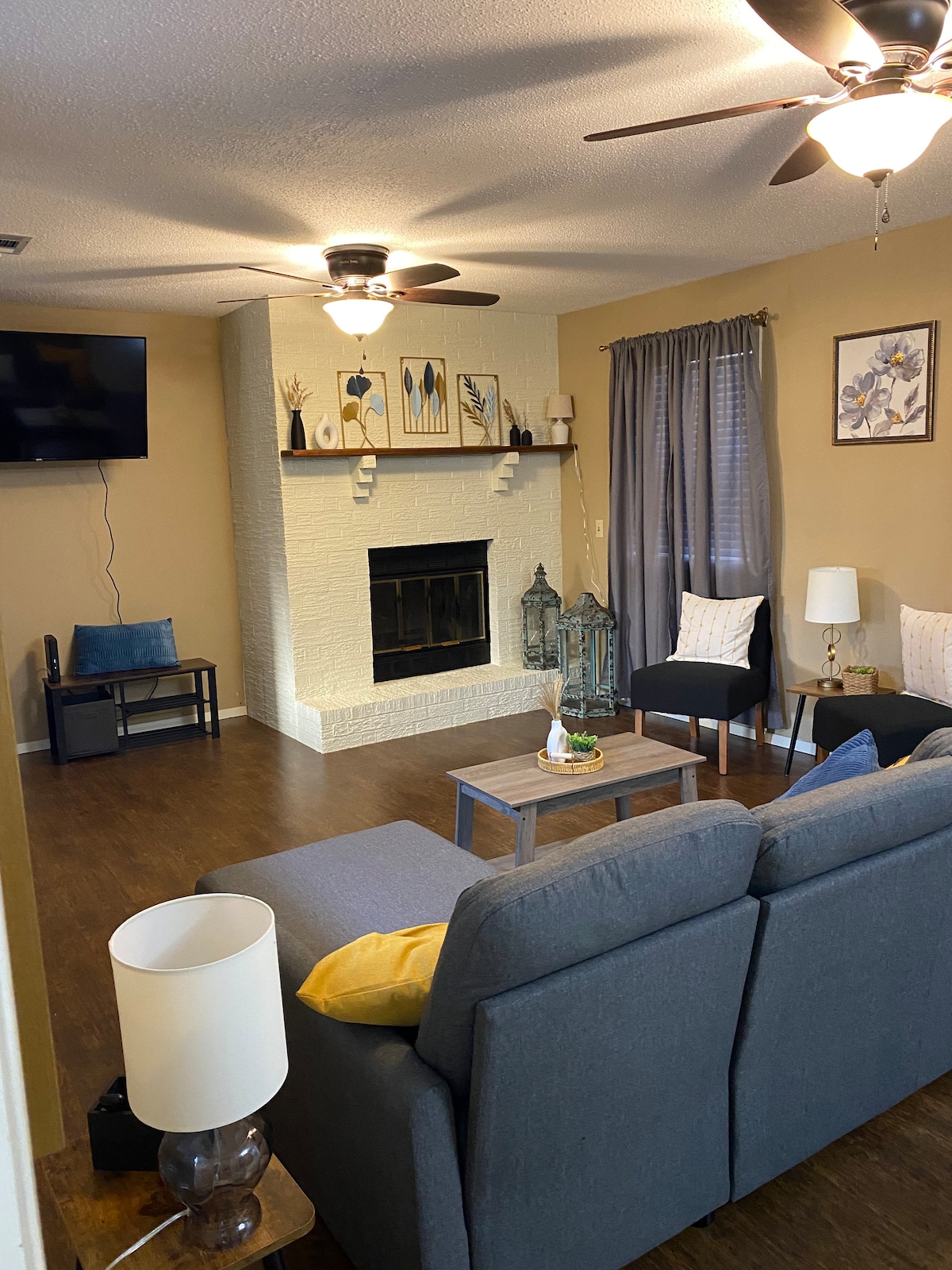 A comfortable living room features a gray sectional sofa, positioned to invite relaxation. A brick fireplace is highlighted by decorative items, while soft lighting from a nearby lamp and ceiling fan create a welcoming ambiance. Two accent chairs and a coffee table complete the cozy setup.