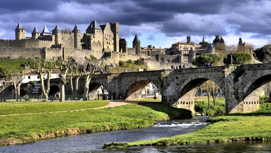 A historic fortress is shown on a hill, featuring multiple towers and a stone wall. A bridge arches over a flowing river in the foreground, with green grass and trees lining the banks. Clouds drift above, creating an atmospheric backdrop.