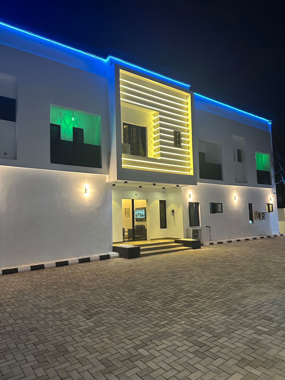 The exterior of the building features a sleek modern design illuminated by multicolored lights at night. The facade is predominantly white, with accents of light creating a striking visual. Several windows provide glimpses into the interior, framed by well-maintained landscaping.