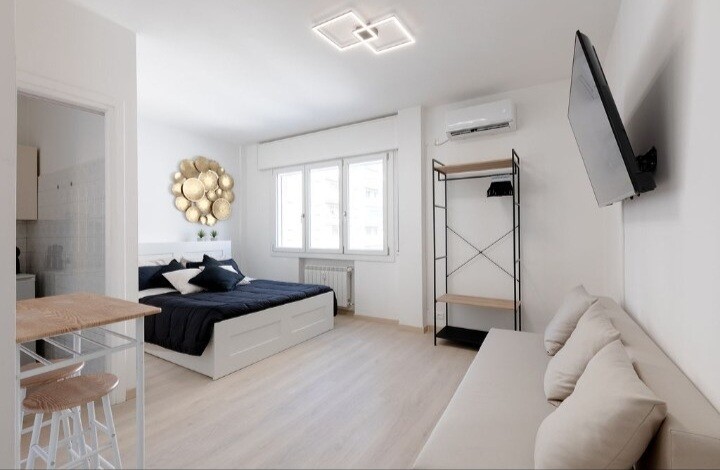 A neatly arranged studio is presented, featuring a spacious bed with dark bedding and decorative pillows. A light-colored sofa complements the wooden shelving unit and table. Bright natural light filters through large windows, emphasizing the room's modern decor and minimalistic style.
