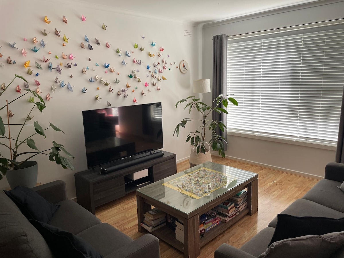 A cozy living area features a television on a low wooden media unit, surrounded by potted plants. A colorful display of origami cranes adorns the wall above. A coffee table with a glass top showcases a puzzle, while natural light filters through the window adorned with blinds.
