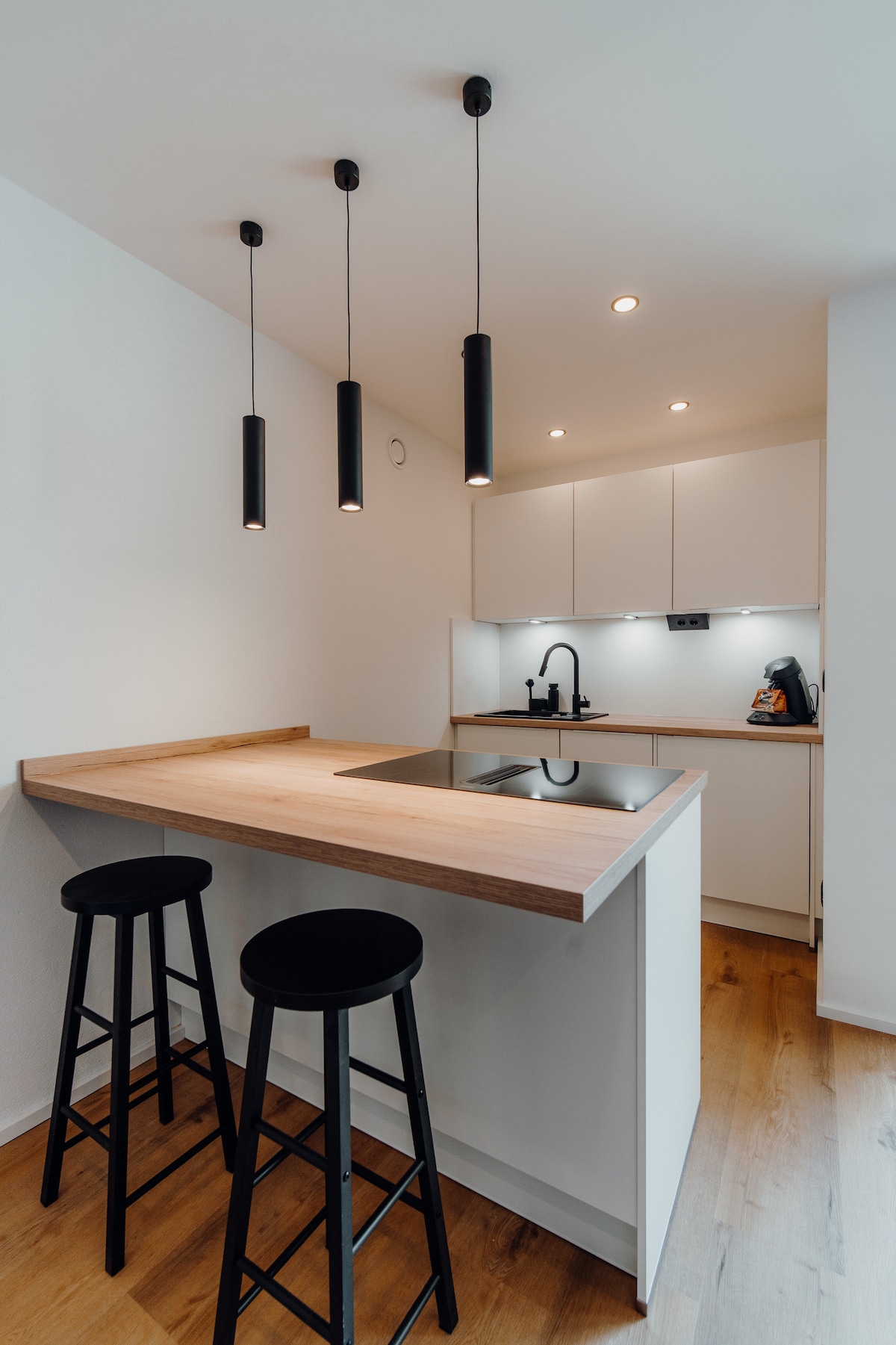 A modern kitchen area is featured, highlighting a sleek countertop with a built-in stovetop. Three black bar stools are positioned at the edge of the counter. Under-cabinet lighting illuminates the space, creating a functional cooking environment. Cabinets provide additional storage options.