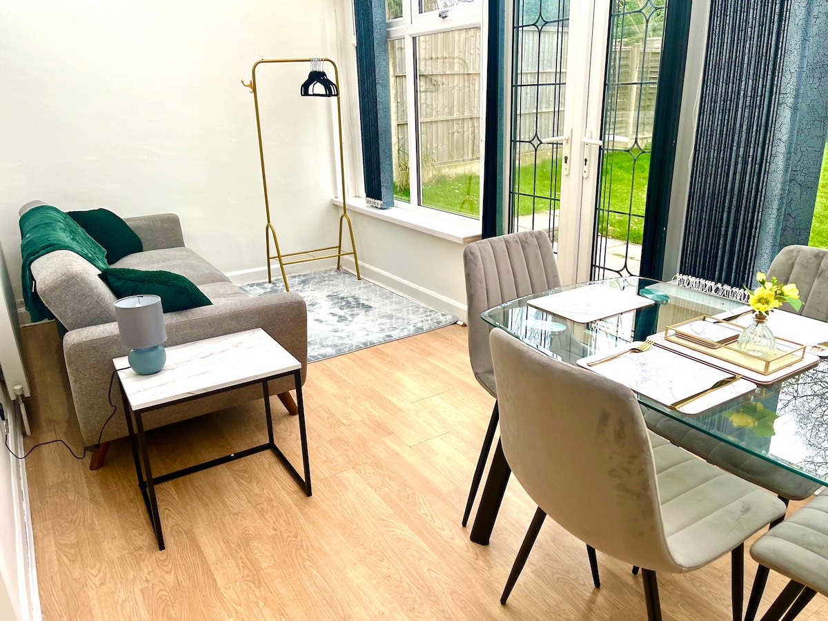 A modern dining area features a glass table set for four with neutral tableware. Plush gray chairs surround the table, and a small side table holds a lamp. A couch with green cushions sits nearby, next to large windows that provide a view of the garden.
