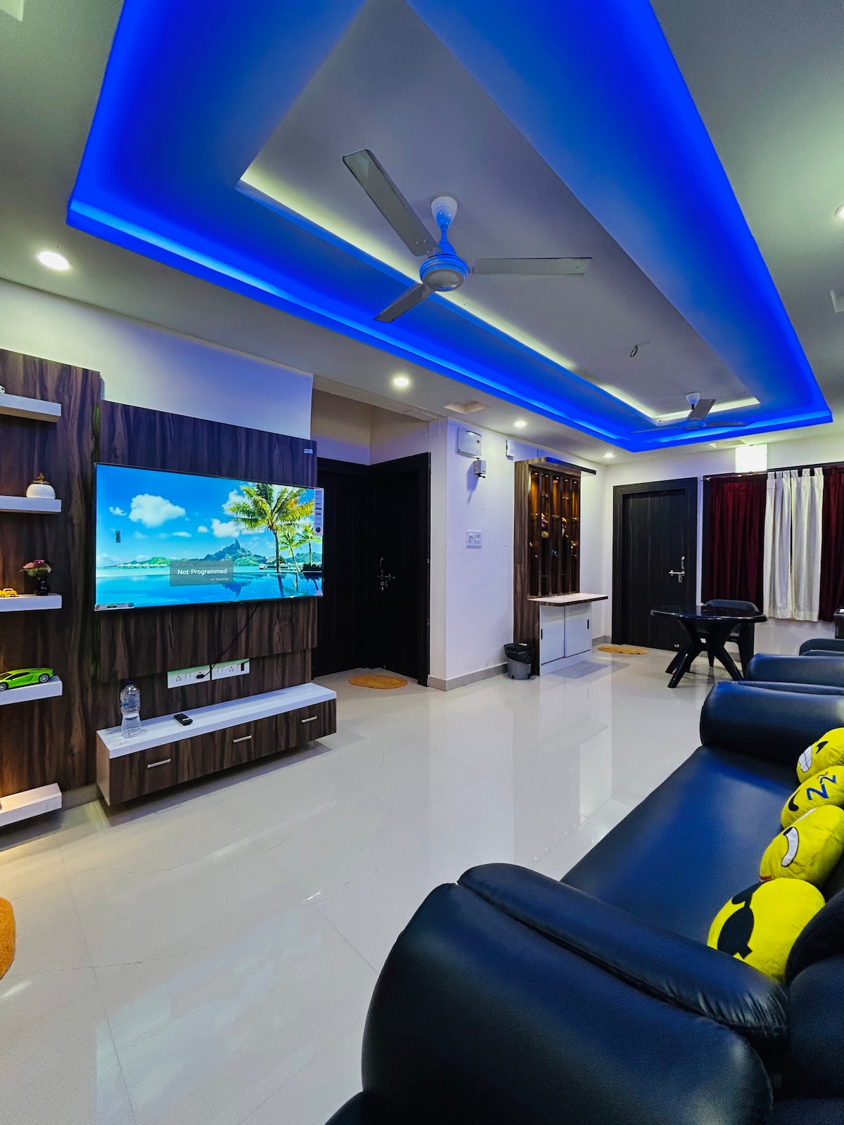 A modern living area features a sleek black sofa with colorful cushions, complemented by ambient blue ceiling lighting. A wall-mounted television displays a scenic beach landscape. Light-colored tiles contribute to a bright environment, while dark wooden accents are visible on the shelves and entertainment unit.