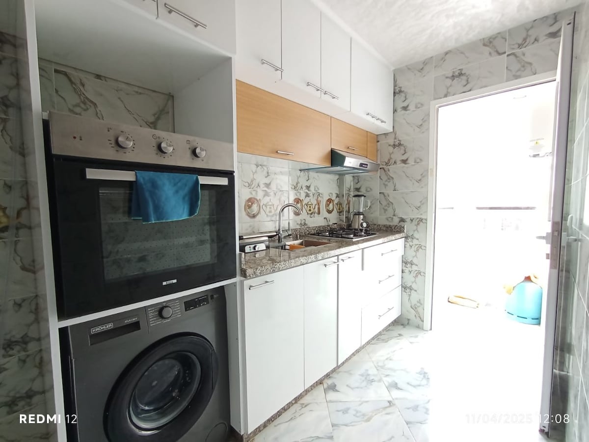 The modern kitchen features a combination of sleek cabinetry and marble-style walls, equipped with a built-in oven and stovetop. A washing machine is positioned next to the kitchen counter, which includes a sink and several appliances. Natural light enters from an adjacent door.