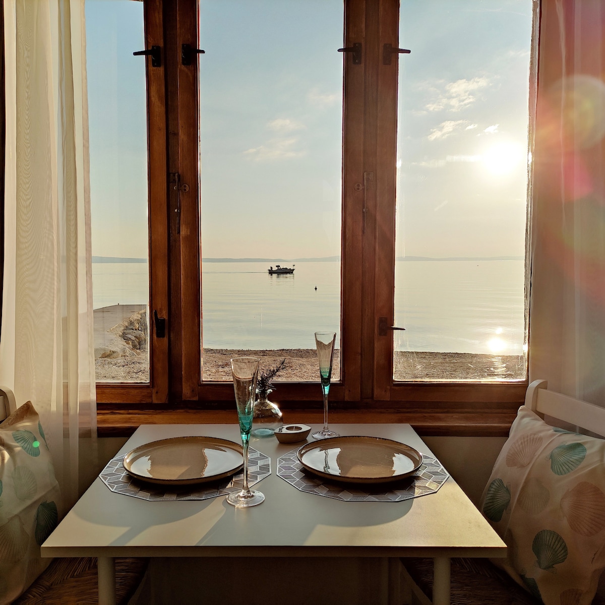 A table set for two is positioned next to large, wooden windows, offering a view of the serene beach and calm waters. Sunlight softly illuminates the space, creating a warm ambiance. Two glasses and plates are neatly arranged on a light-colored table, inviting guests to enjoy a meal.
