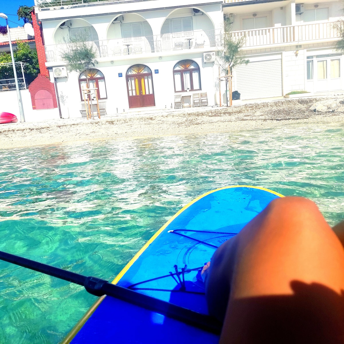A clear view of calm waters reflects the surrounding building, featuring large arched windows and a welcoming terrace. A paddleboard is partially visible in the foreground, with a person's leg resting on it, indicating the proximity to water activities.
