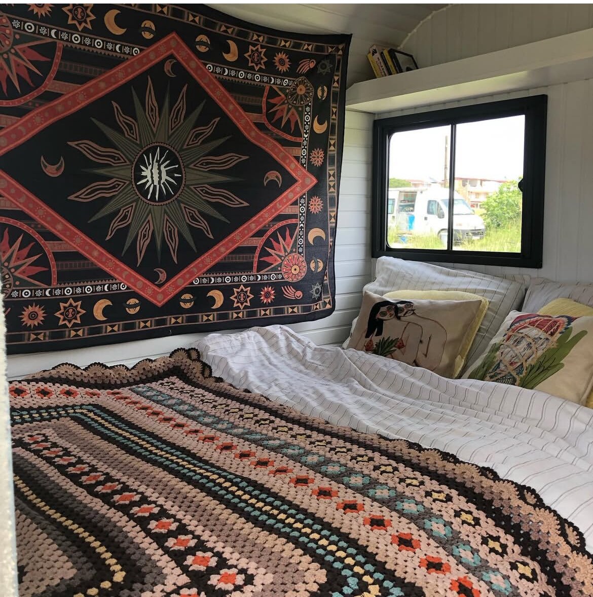 A cozy sleeping area is presented with a large, colorful tapestry on the wall, complemented by decorative cushions. A spacious bed is adorned with a patterned blanket, and light filters in through a nearby window, illuminating the inviting interior of the caravan.