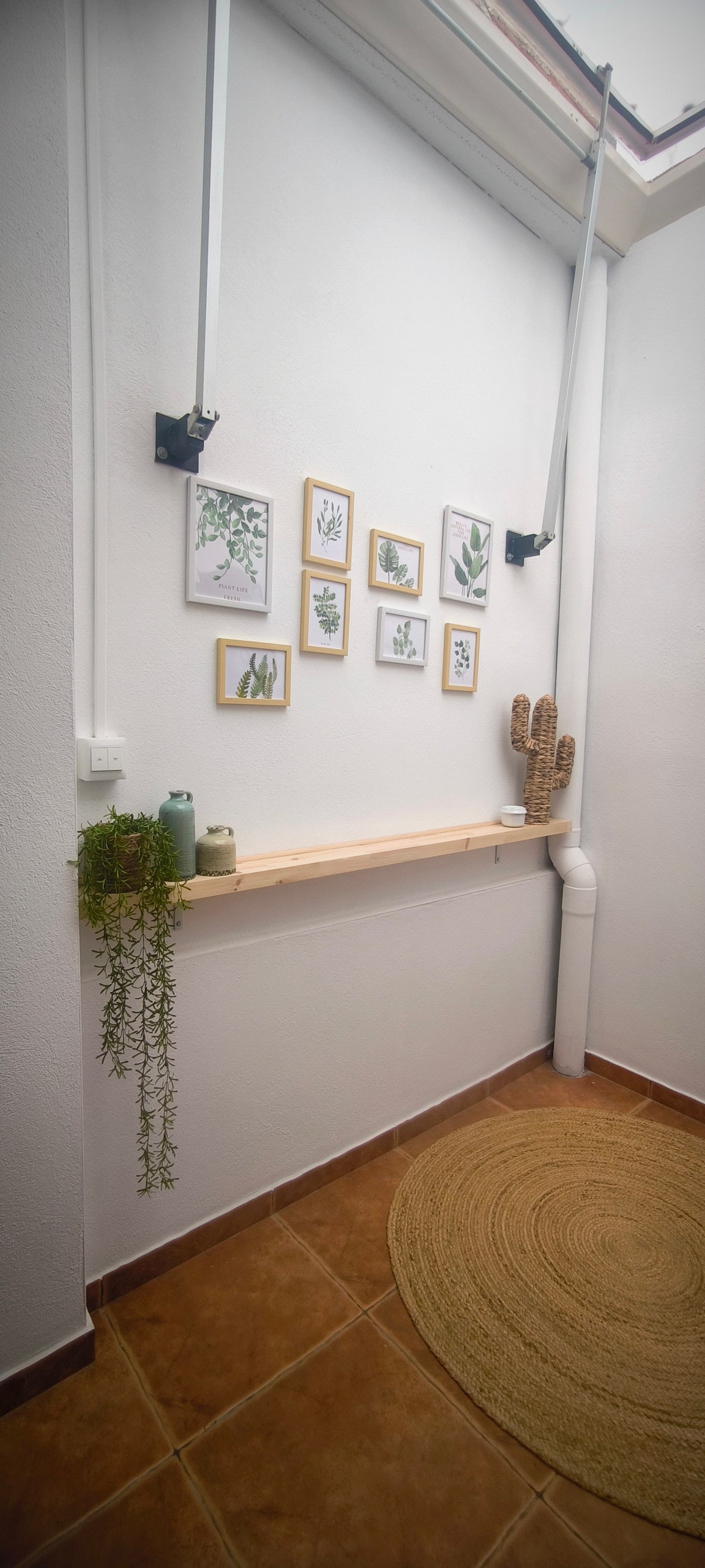 A narrow hallway features white walls accented by a series of botanical art prints in wooden frames. A wooden shelf holds several small green plants, while a round, natural fiber rug lies on the tiled floor, creating a warm and inviting space.