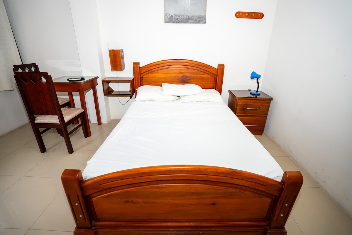 A bedroom is featured with a wooden bed and a simple white bedding. A wooden nightstand holds a blue lamp, while a matching chair and table are situated nearby. Natural light enters through a window, adding brightness to the neutral-toned room.
