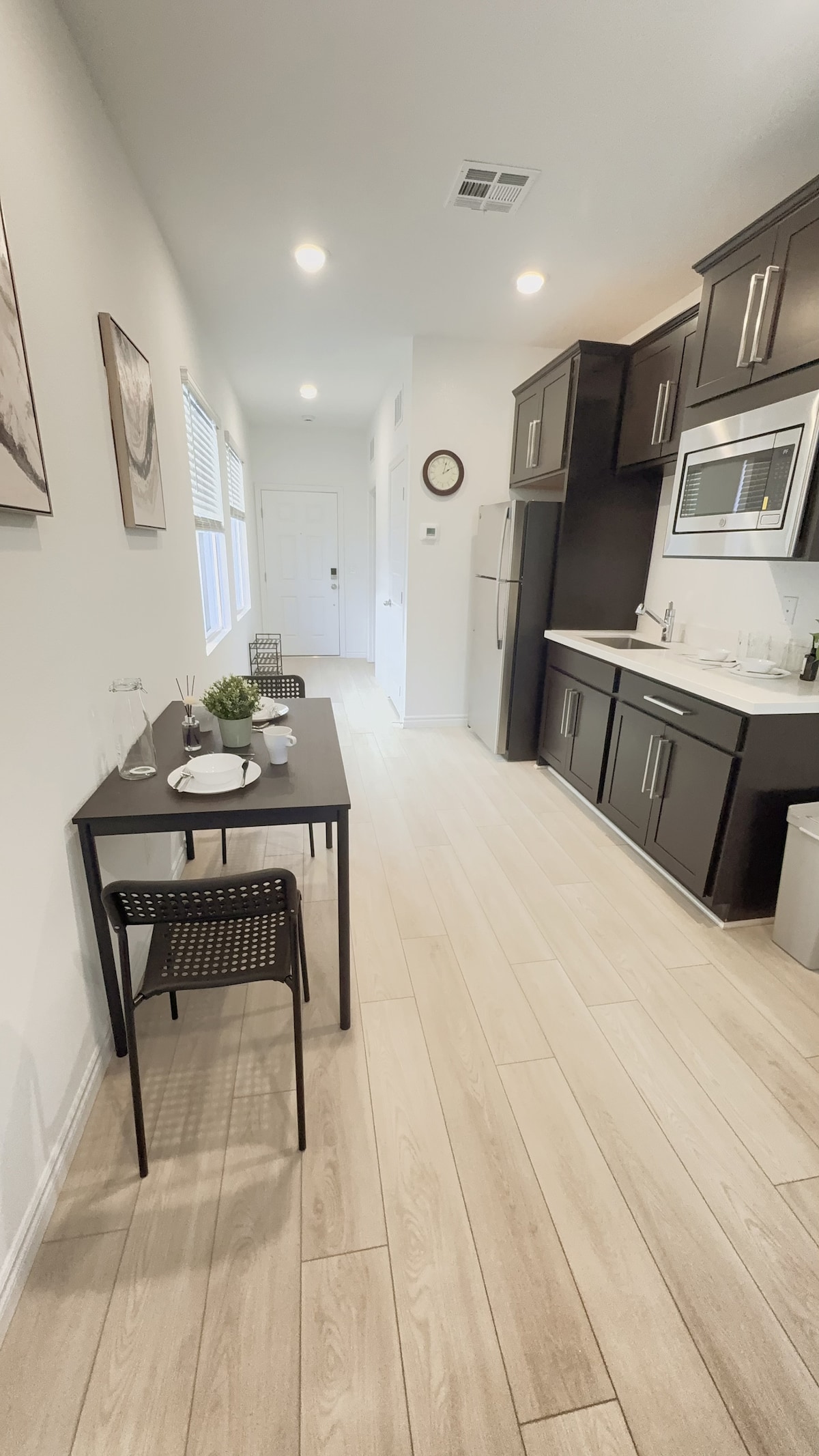 The kitchenette features dark cabinetry and modern appliances, including a microwave and refrigerator. A small dining table with two chairs is positioned beside a window, allowing natural light. The open space leads to a private entrance with a light-colored floor.