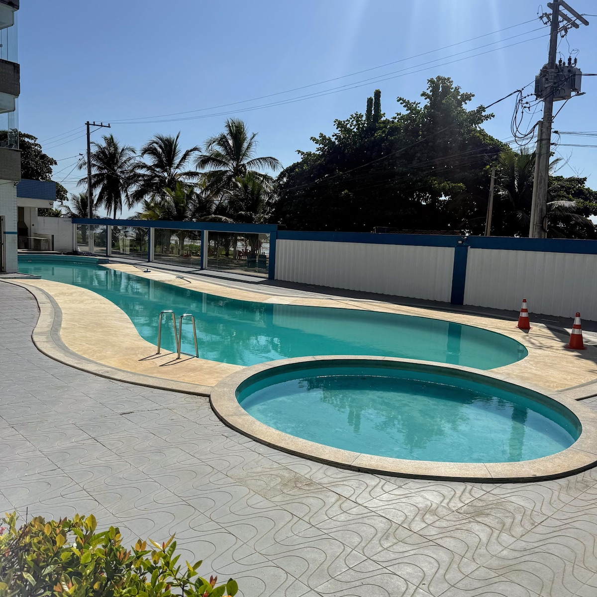 A tranquil pool area is visible, featuring a long rectangular pool alongside a smaller circular pool. Palm trees provide shade around the space, while clear blue water reflects the sunlight. The pool deck is bordered by a paved walkway, with safety cones placed nearby.