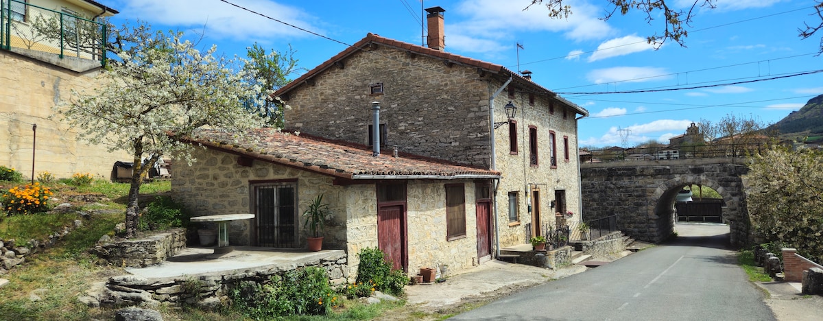 A rustic stone house is presented along a quiet road, featuring a tiled roof and wooden shutters. A small terrace with seating is visible on the left, while an archway bridges over the road in the background, framed by greenery and colorful flowers blooming nearby.