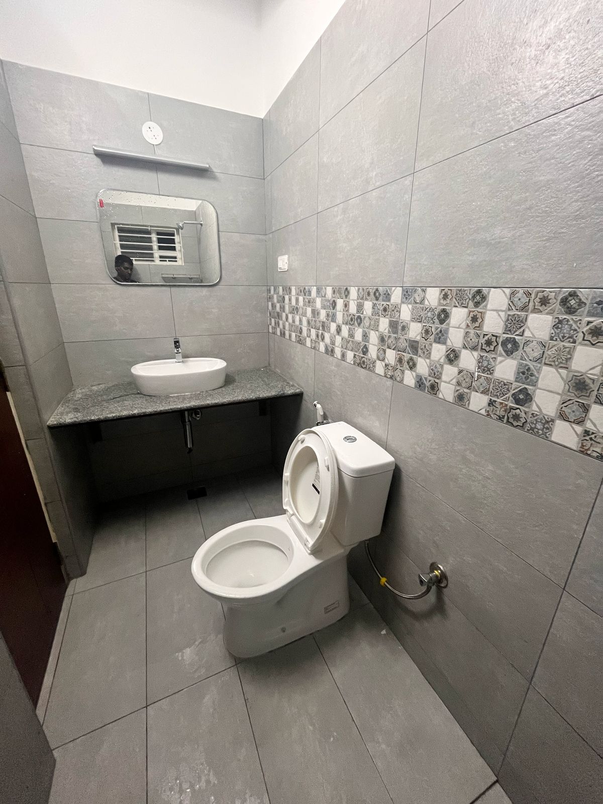 The bathroom features grey tiled walls and floors, complemented by a stylish sink with a wall-mounted mirror above it. A modern toilet is positioned discreetly, while a patterned tile border runs along the wall, adding a detailed touch to the design.