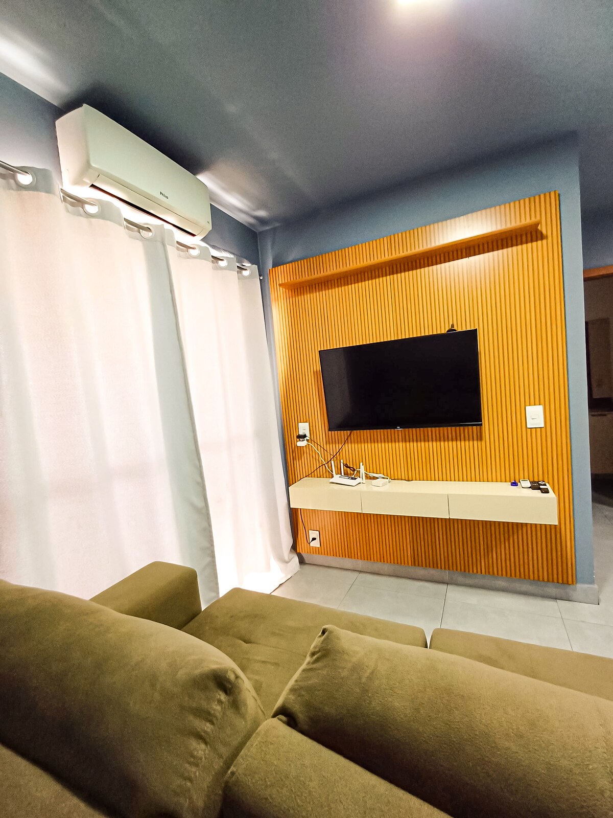 A comfortable seating area is presented with a plush sofa positioned in front of a wall-mounted television. Air conditioning is visible above, and sheer white curtains softly diffuse natural light. The wall features a warm wooden accent for added texture.