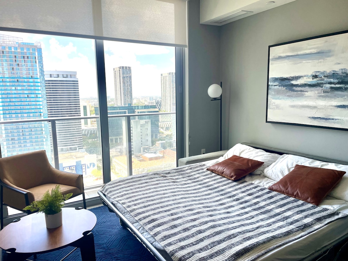 A modern sleeping area features a spacious sofa bed with decorative cushions, complemented by a stylish armchair nearby. Large windows provide a panoramic view of the city skyline, with soft natural light illuminating the room. A small table with a potted plant adds a touch of greenery.