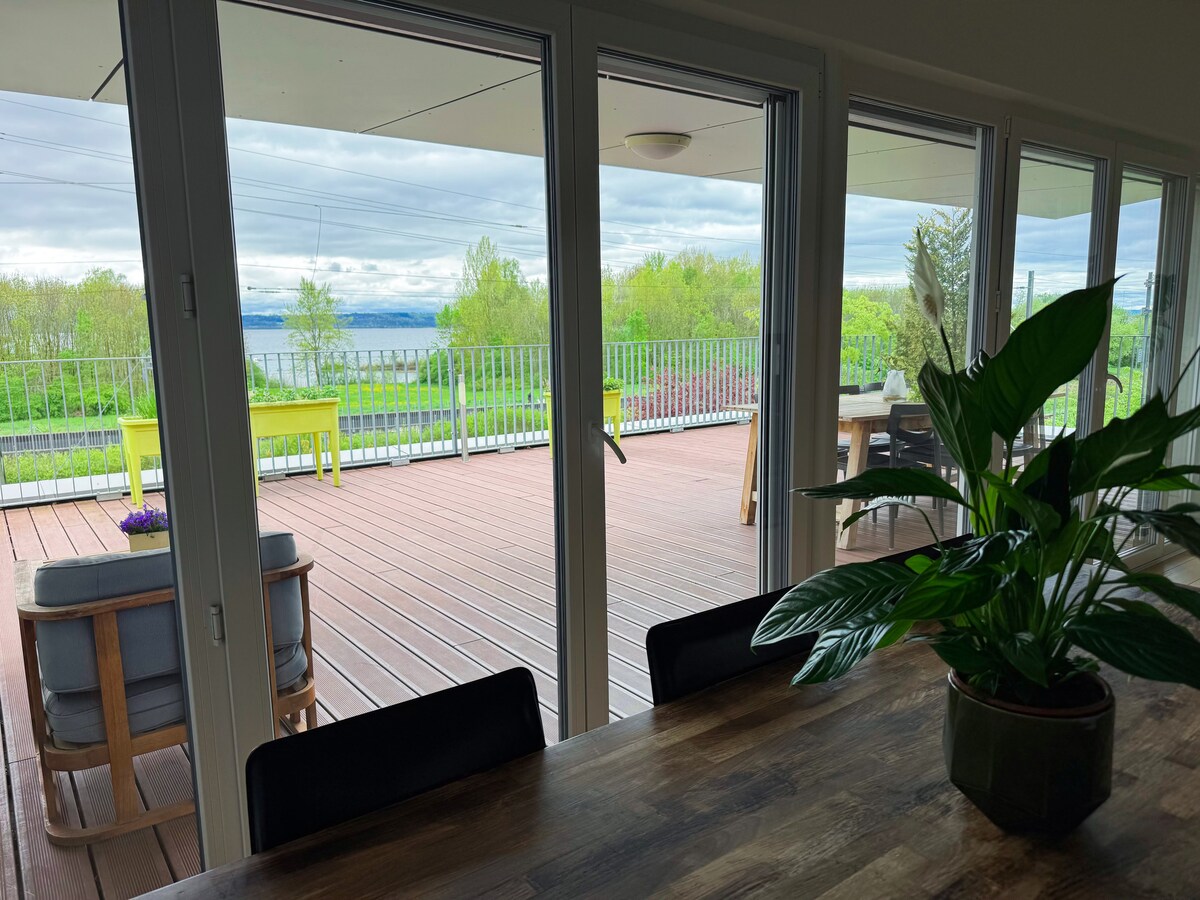 Large glass doors open onto a spacious terrace, featuring a scenic view of green surroundings and the lake in the distance. A dining area is visible indoors, complemented by a potted plant on the table and comfortable seating arranged on the terrace.