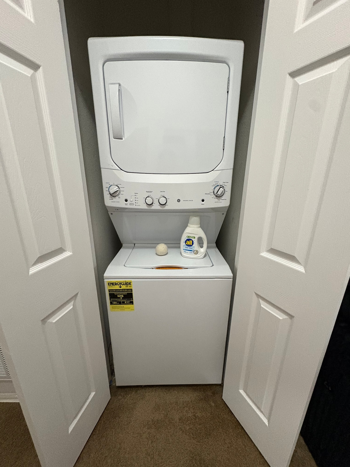 A stacked washer and dryer are situated within a narrow closet. The washer is located at the bottom, topped by the dryer. Laundry detergent is placed on the dryer. The closet doors are open, revealing the appliances in a neutral-toned space.