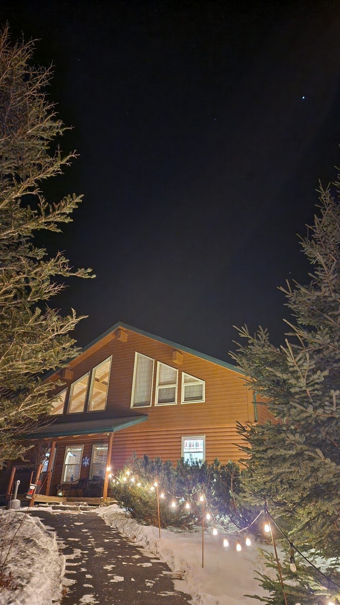 The exterior of a wooden house is illuminated by soft pathway lights, creating a warm presence amidst a snowy landscape. Tall trees frame the path leading to the entrance, which features large windows revealing the interior's welcoming ambiance.