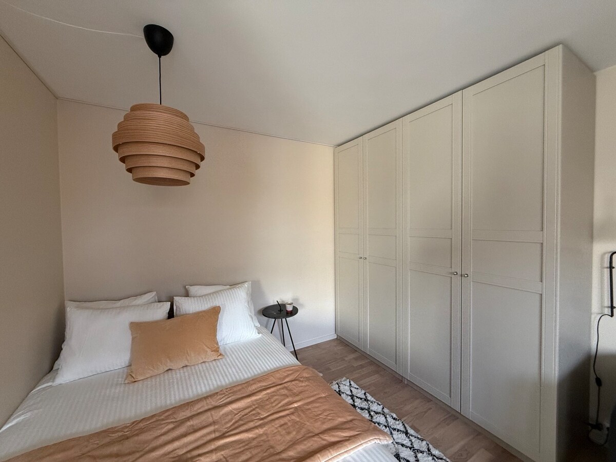 A cozy sleeping area features a spacious bed with neatly arranged white linens and an accent pillow. A stylish pendant light hangs above, and two large wardrobes are visible against the wall. A small side table with a candle completes the warm ambiance.