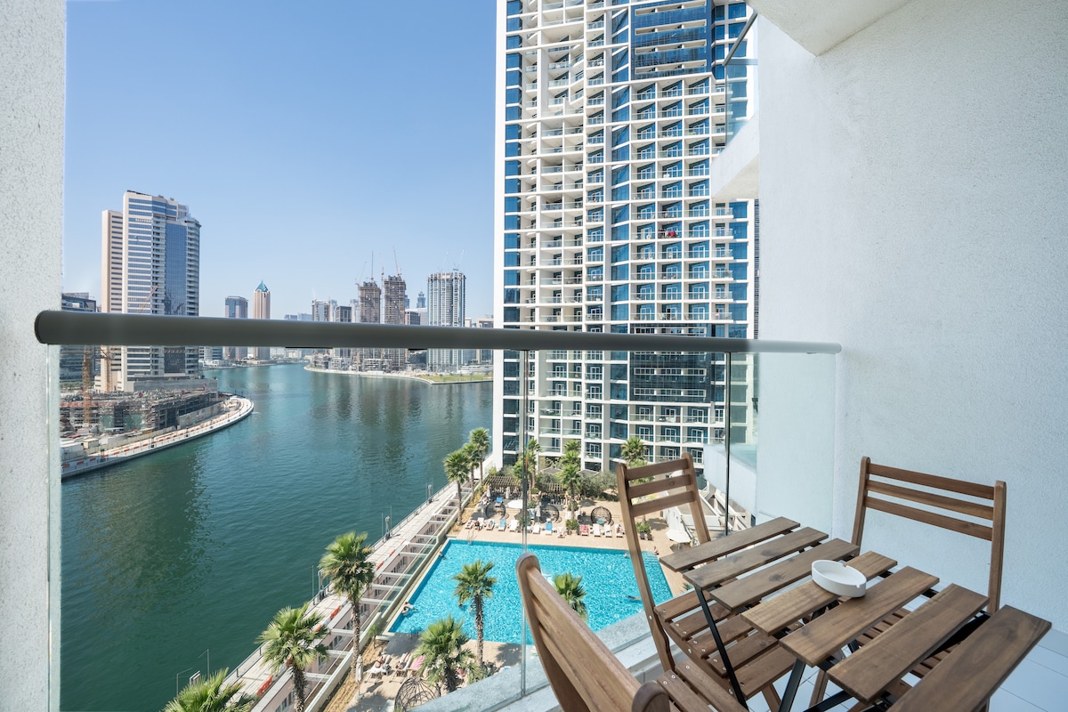 A balcony view reveals the sparkling waters of Dubai Canal, with lush palm trees lining the pool area below. The sleek architecture of nearby buildings contributes to the modern cityscape under a clear blue sky.
