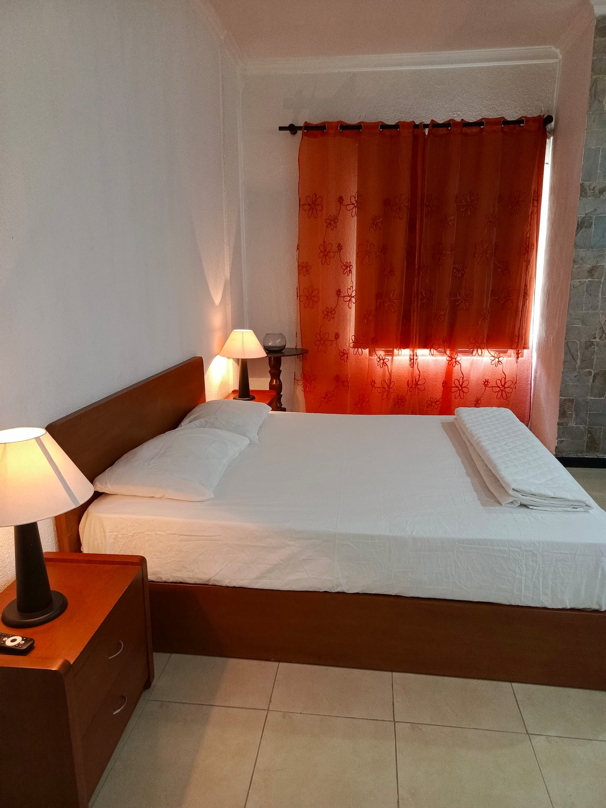 A bright sleeping area features a spacious bed topped with white linens. Soft lighting is provided by two bedside lamps, while a large window is dressed with sheer orange curtains, allowing natural light to filter through. A wooden nightstand complements the warm tones of the decor.