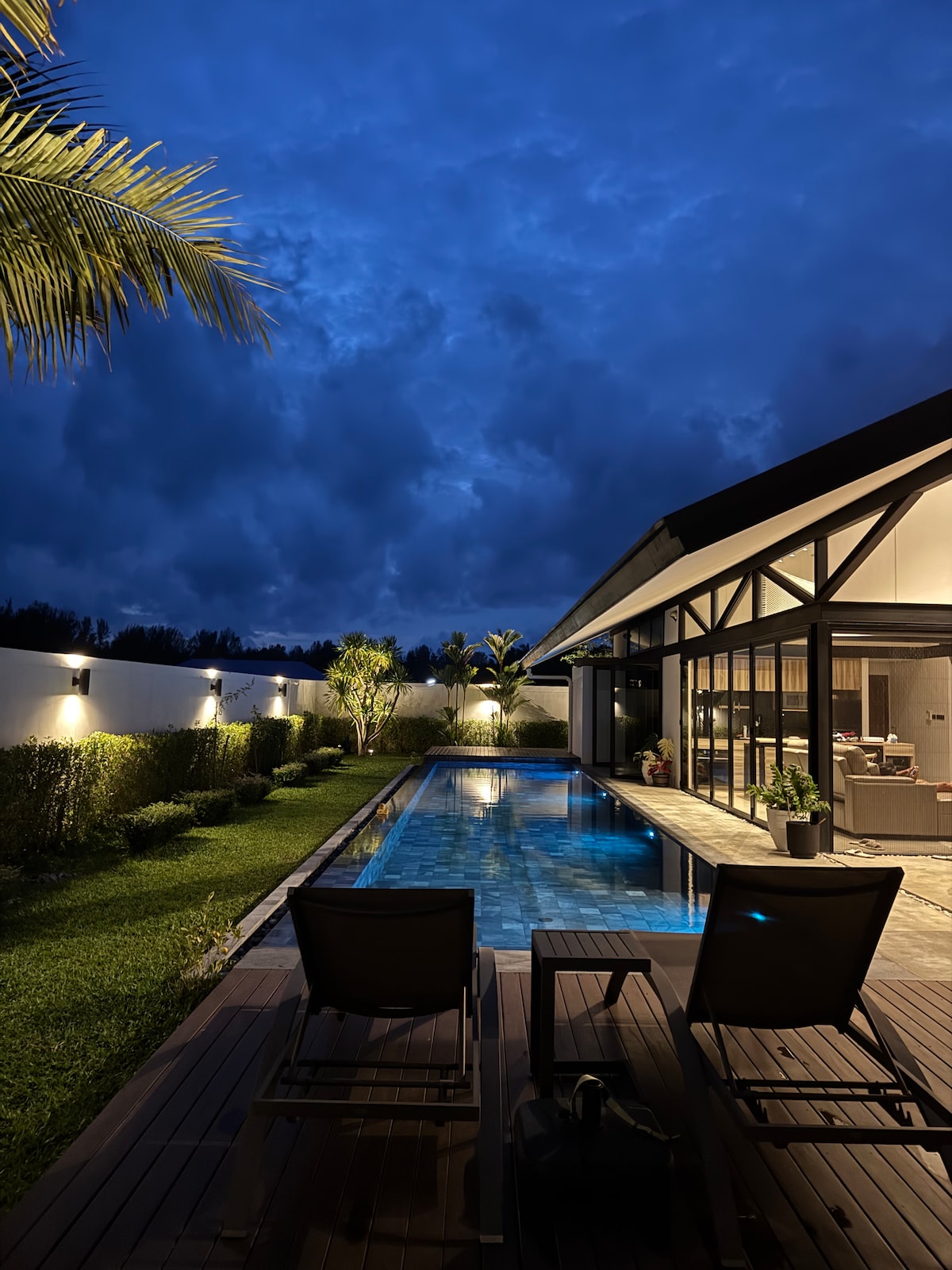 A serene view of a private pool is presented, surrounded by lush greenery and well-maintained gardens. Two sun loungers are positioned beside the pool, inviting relaxation. The night sky is filled with dramatic clouds, and soft lighting illuminates the villa's exterior, enhancing the tranquil atmosphere.