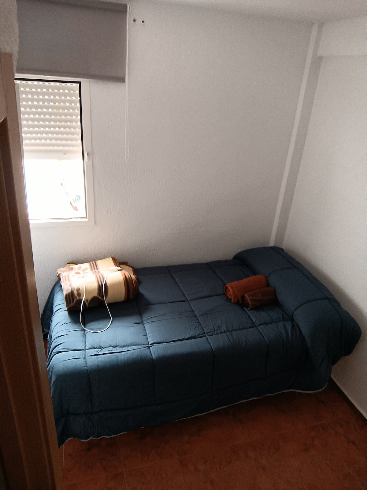 A small sleeping area features a single bed covered with a blue quilt. A window allows natural light to fill the space, complemented by a neatly arranged set of towels at the foot of the bed. The room is characterized by its minimalist decor.