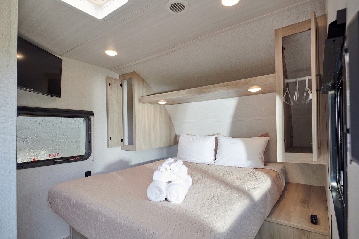 The sleeping area features a spacious bed with a neutral bedspread and neatly arranged towels. Ambient lighting is provided by ceiling fixtures, and storage cabinets are integrated into the wall. A small television is mounted on the wall opposite a window, providing natural light.