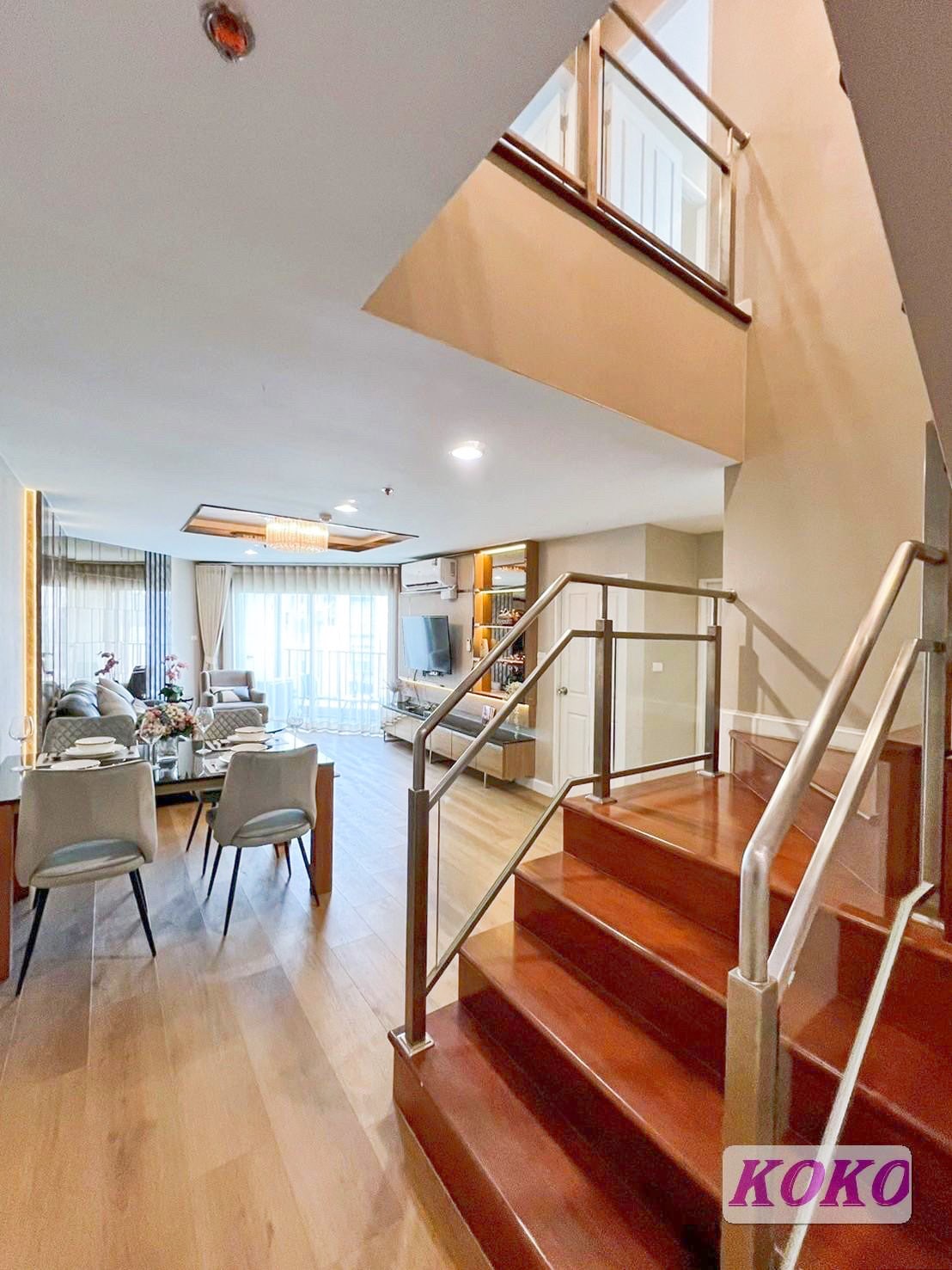 A staircase with polished wooden steps is featured, leading to an upper level. The light-filled living area can be seen in the background, highlighting a modern design with comfortable seating and large windows adorned with soft curtains.
