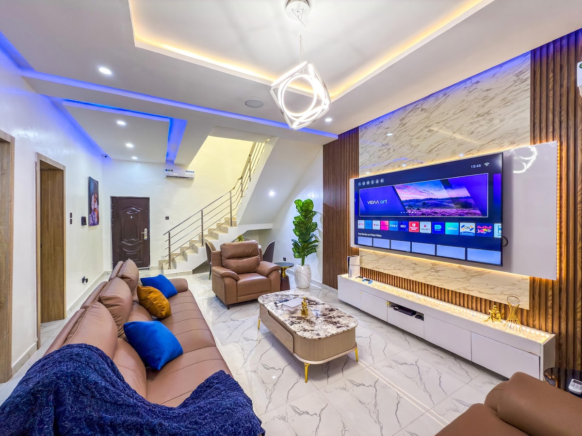 A spacious living area is highlighted by a large mounted television and contemporary furniture. Soft brown sofas are arranged around a stylish coffee table. Soft, blue lighting enhances the modern decor, while a staircase is visible in the background, leading to the upper level.