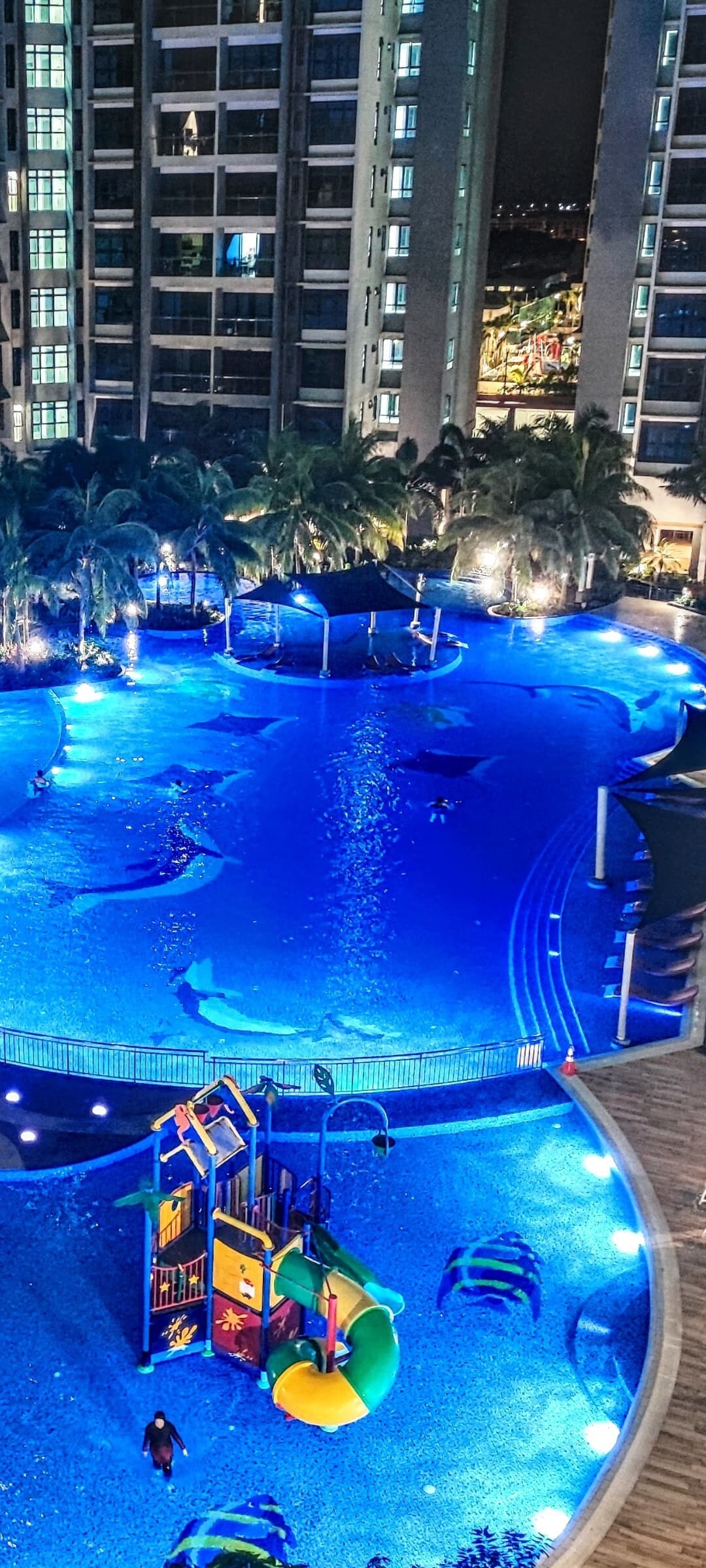 A well-lit swimming pool is surrounded by palm trees, reflecting blue hues under the night sky. A colorful playground is positioned nearby, featuring slides and climbing structures, adding a playful element to the serene environment.
