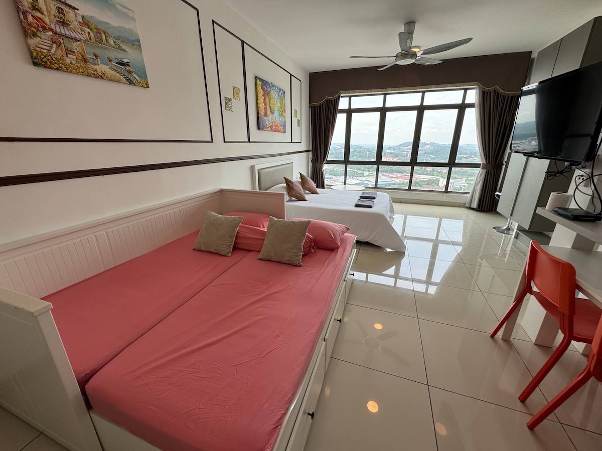 A spacious studio unit features a pink sofa bed and a comfortable queen size bed. Large windows allow natural light to flood the room, offering a panoramic view of the surroundings. A television is mounted on the wall opposite the beds, completing the cozy living space.