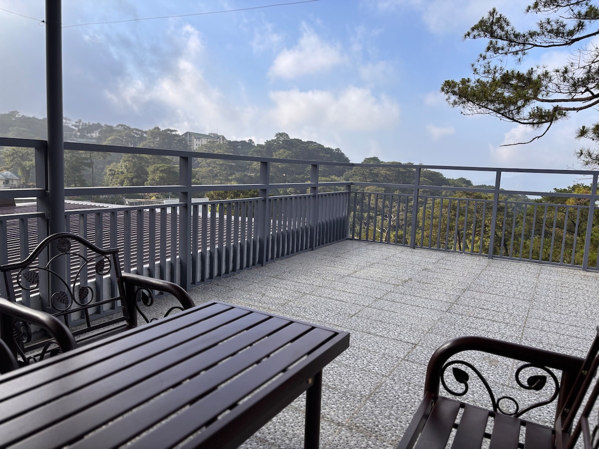 An outdoor terrace is featured, showcasing a wooden table accompanied by two matching chairs. The space is surrounded by a railing with views of lush greenery and gentle hills under a brightened sky, creating a serene spot for relaxation.