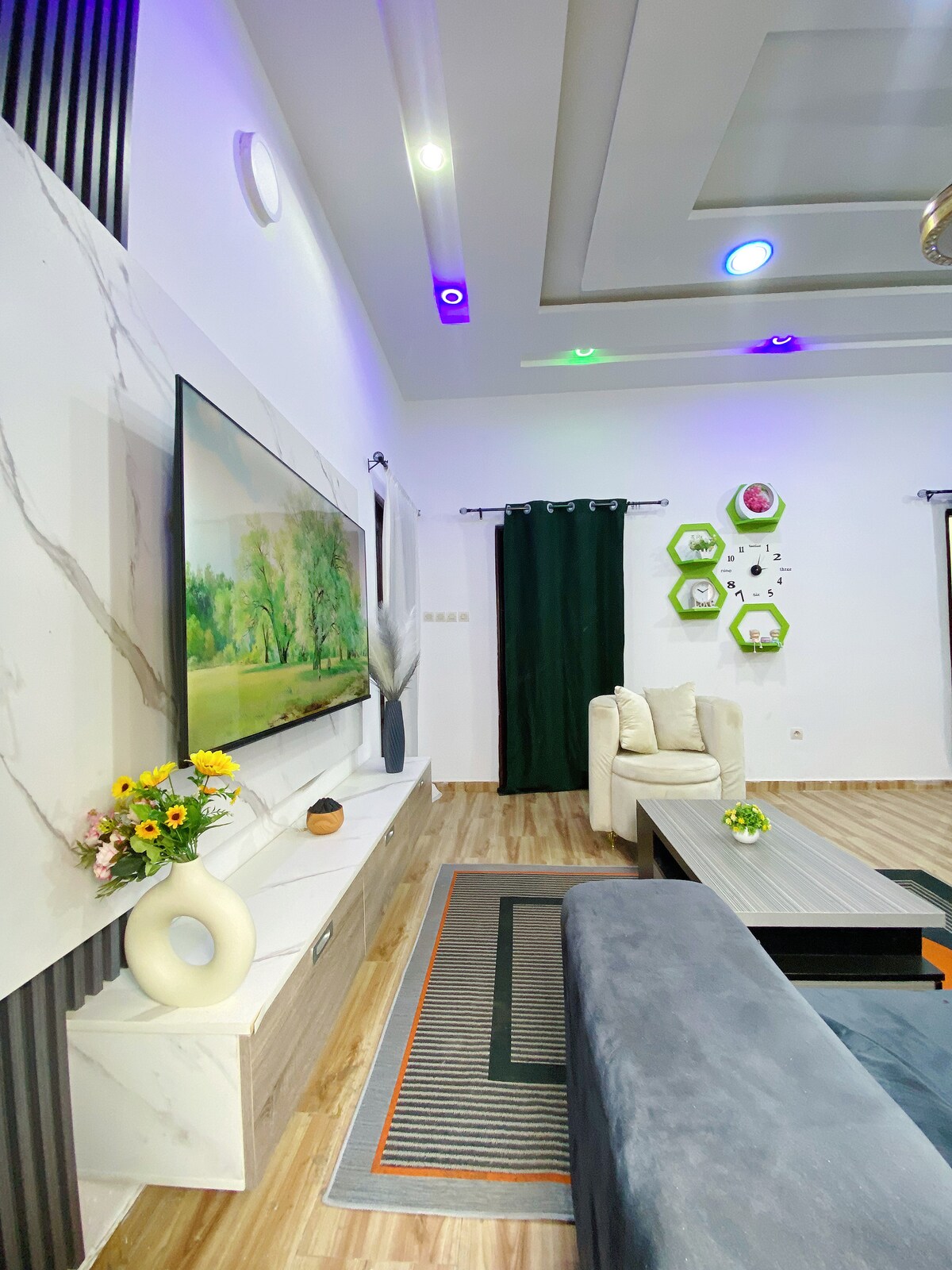 A modern living area features a large wall-mounted TV displaying a landscape scene. A light-colored sectional sofa complements a neutral coffee table, which is adorned with a small plant. Decorative elements include colorful wall accents and curtains framing a window.