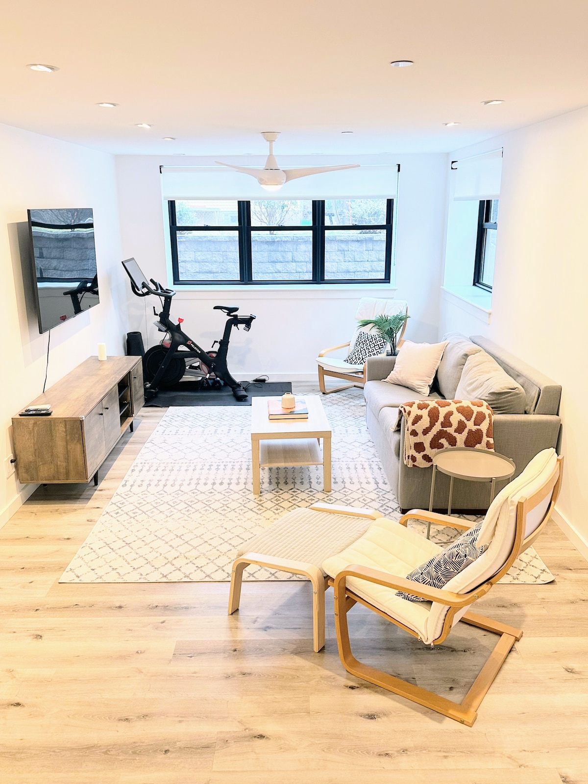A modern living area features a grey sofa paired with a decorative throw and cushions. A small coffee table is positioned in front, with a light area rug underfoot. A stationary bike stands beside the TV, which is mounted on the wall, and large windows allow natural light to enter.