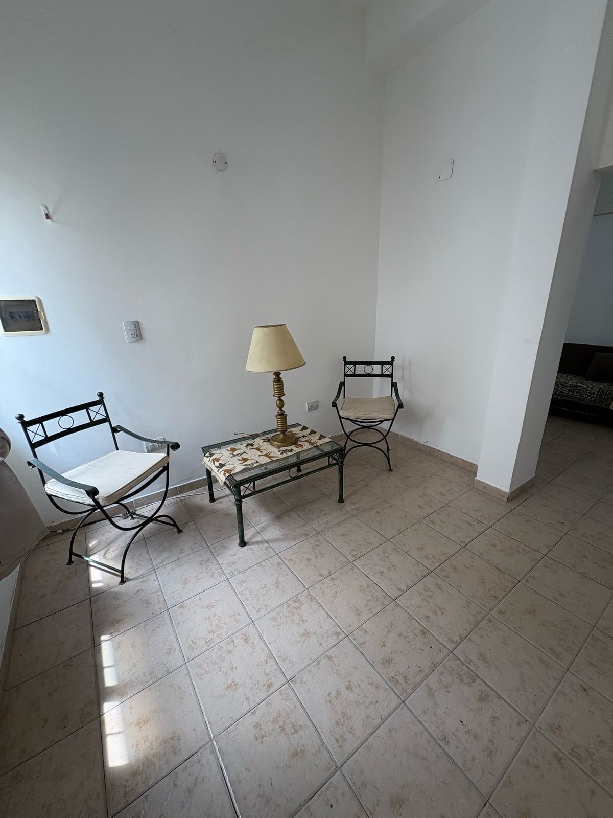 A corner of the living area features two metal chairs positioned around a small, decorative table. A standing lamp provides lighting. Sunlight illuminates the tiled floor, enhancing the spaciousness of the neutral-colored walls.