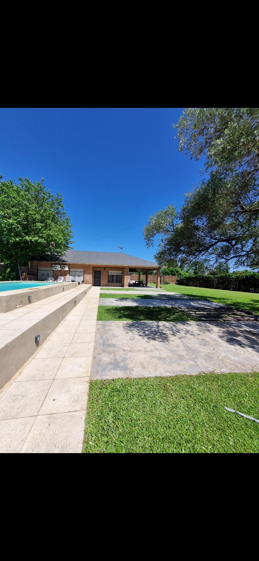 A spacious outdoor area is observed, featuring a well-maintained lawn and a stone pathway leading towards a single-story building. The structure is surrounded by greenery, with a large tree casting shade. A pool is visible in the foreground, reflecting the clear blue sky.