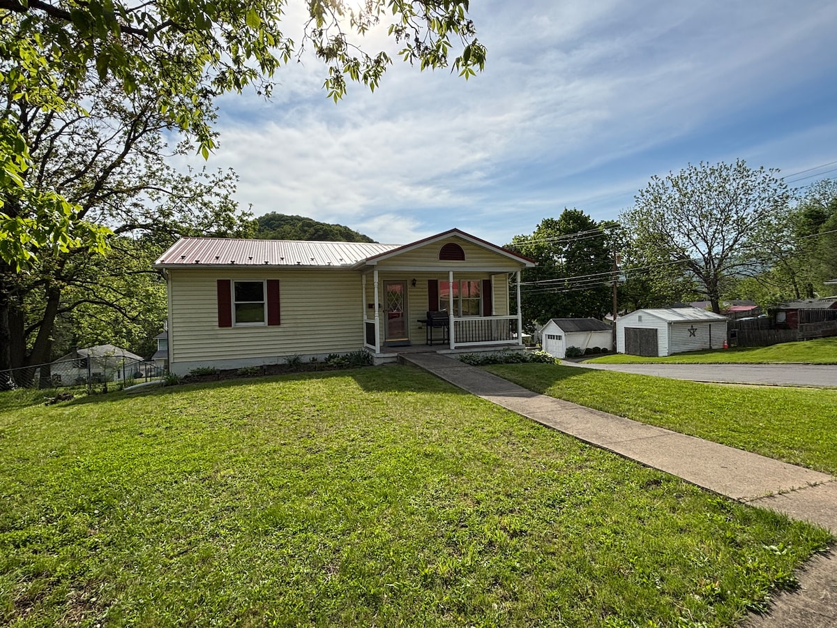 A single-story home is set on a grassy lot, framed by mature trees and shrubs. A concrete path leads to a covered porch showcasing a white rocking chair. The house features a light-colored exterior with red accents and a metal roof, providing a welcoming appearance.