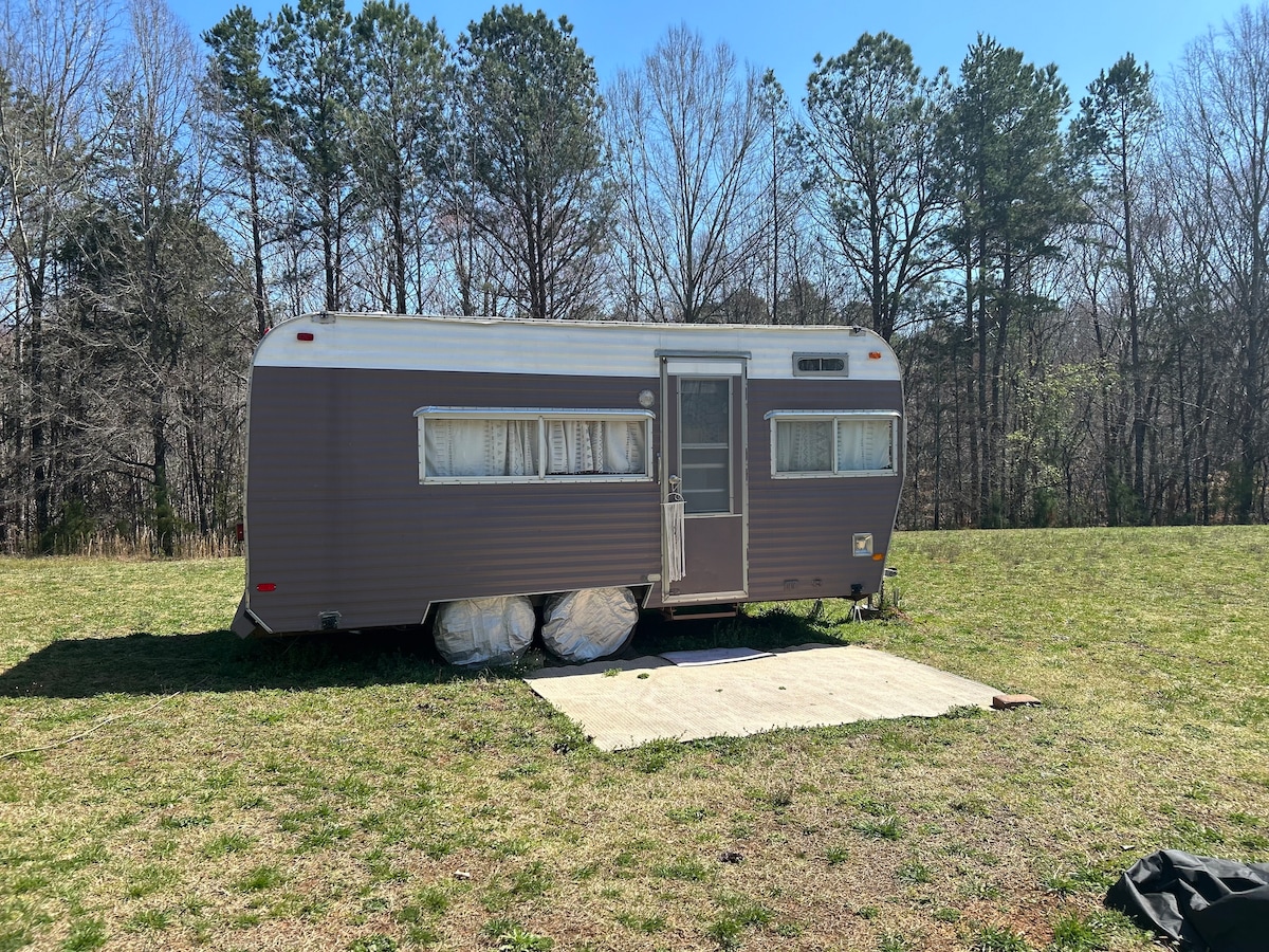 A vintage travel trailer is positioned on a grassy area, surrounded by trees in the background. The trailer features a light brown exterior with large windows, partially covered by sheer curtains. A concrete slab serves as the foundation beneath the trailer.