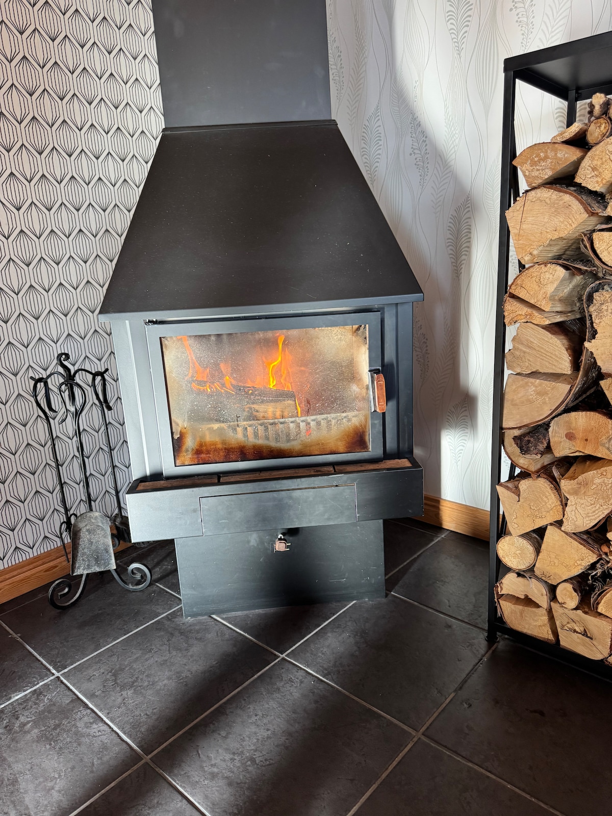 A modern fireplace is featured, with flames visible through the glass door. It is set in a corner beside a stack of neatly arranged firewood. The surrounding area is tiled, complementing the contemporary design of the home.