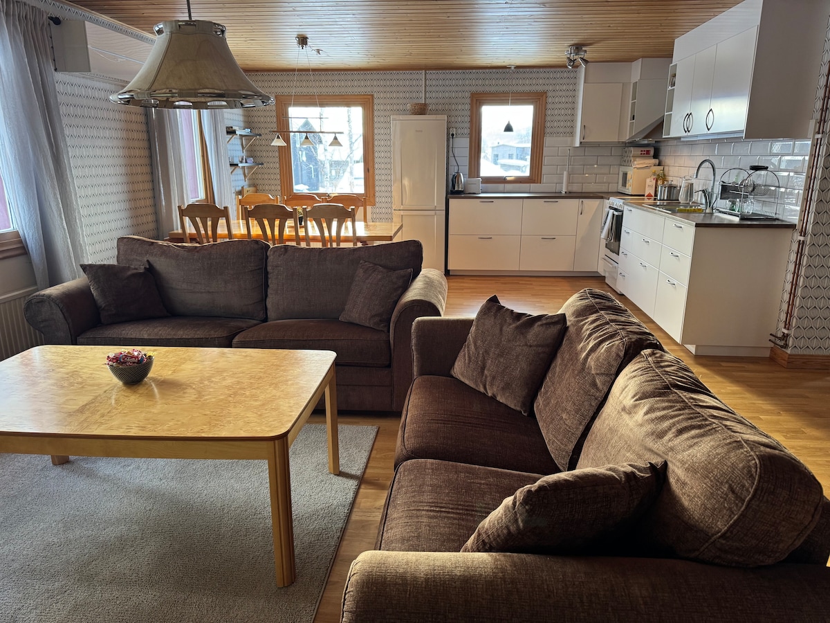 A spacious living area features two comfortable brown sofas arranged around a wooden coffee table. A dining table with chairs is positioned in the background, alongside a fully equipped kitchen. Large windows allow natural light to fill the room, highlighting the warm wooden accents.