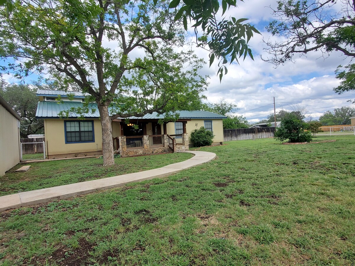A single-story house with a green metal roof is surrounded by a spacious yard, featuring a curved pathway leading to the entrance. Mature trees provide shade alongside the home, while an open area with grass enhances the inviting outdoor space.