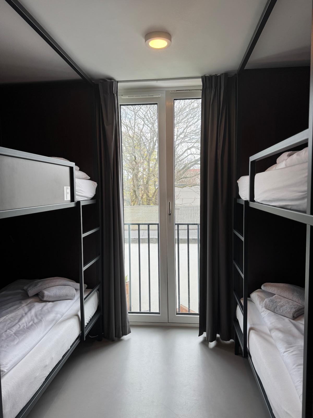 A compact sleeping area features two sets of bunk beds with neatly arranged bedding. Light enters through a large glass door that leads to a balcony, with dark curtains framing the window. The walls are designed in a deep hue, enhancing the cozy feel.