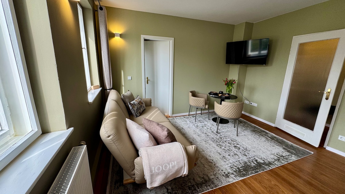 A comfortable living area is captured, featuring a light-toned sofa with decorative pillows. A green accent wall complements the modern furnishings. A flat-screen TV is mounted on the wall, and a round dining table is set with fresh flowers, creating an inviting space.