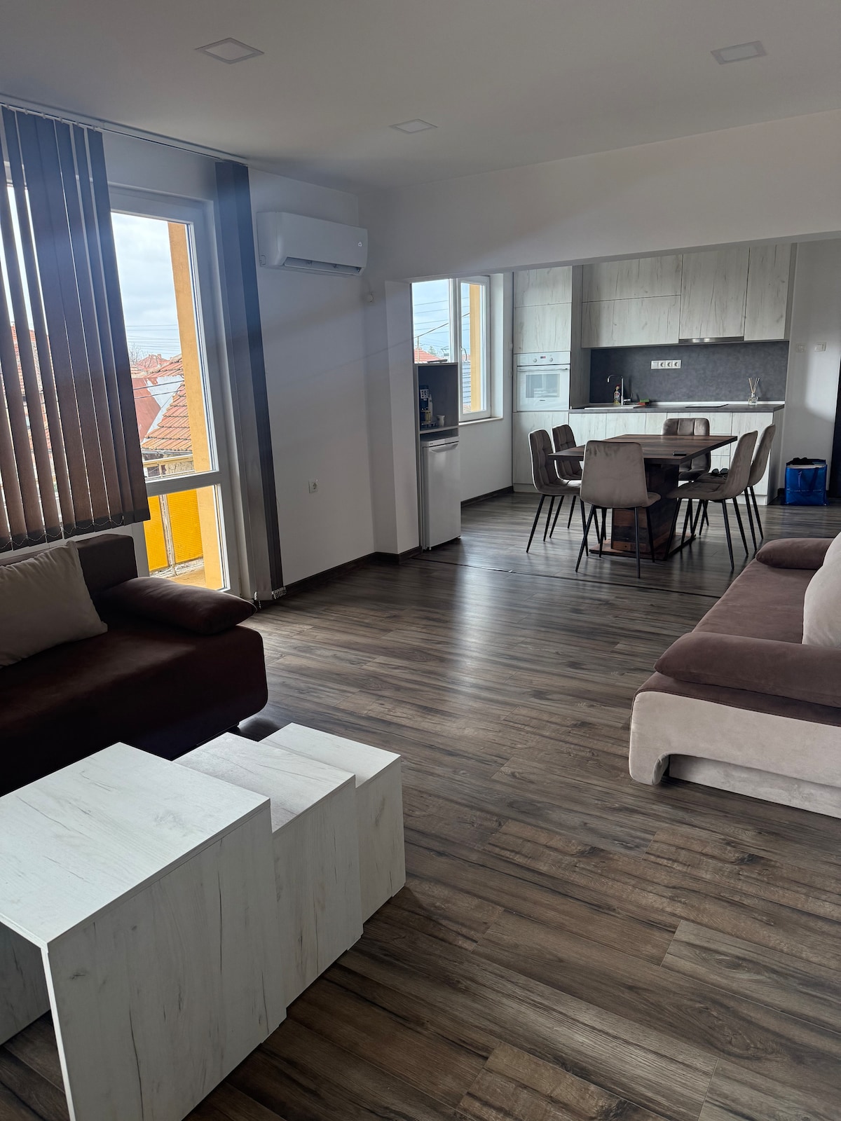A modern living space is highlighted by large windows allowing natural light. A dining area with a table and chairs is visible in the background, alongside a kitchen with contemporary cabinetry. Comfortable sofas are positioned in the foreground, complementing the wooden flooring.