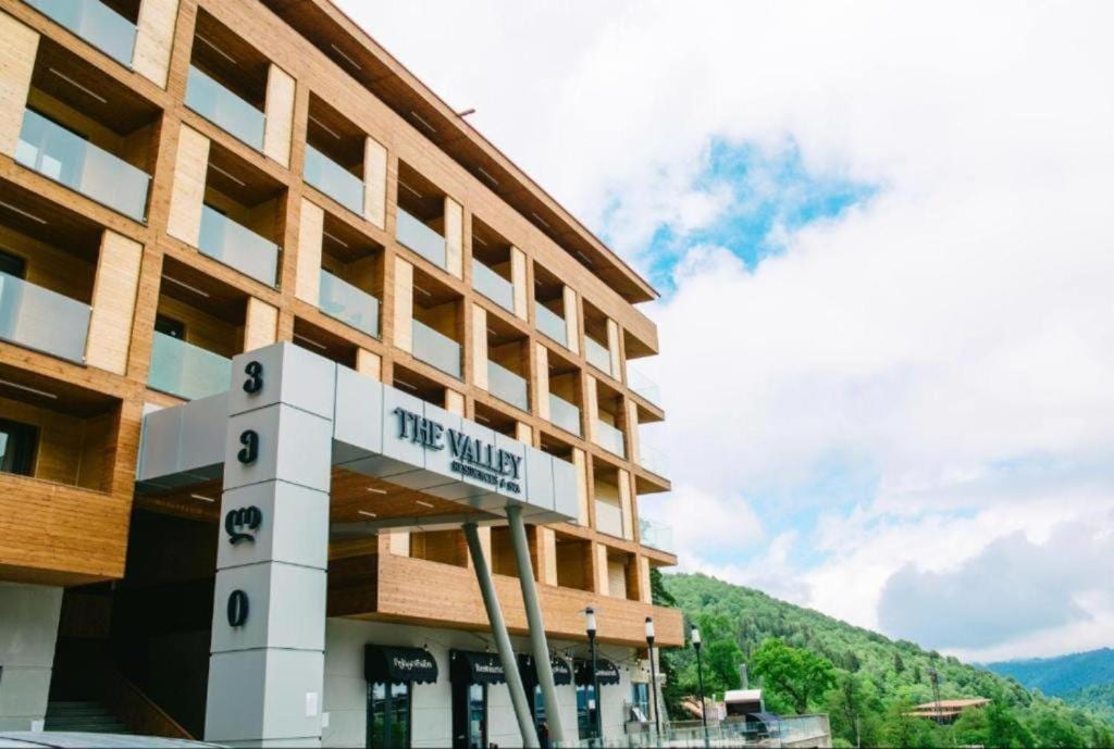 The exterior of The Valley Residence & Spa is captured, showcasing a modern building with a combination of wood and stone finishes. The entrance features large, inviting glass doors. Lush greenery surrounds the property, with a backdrop of rolling hills under a partly cloudy sky.